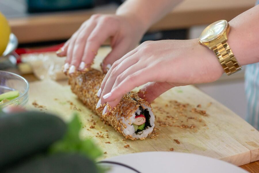 Student hands pressing sesame-seed-rolled sushi on a wooden board